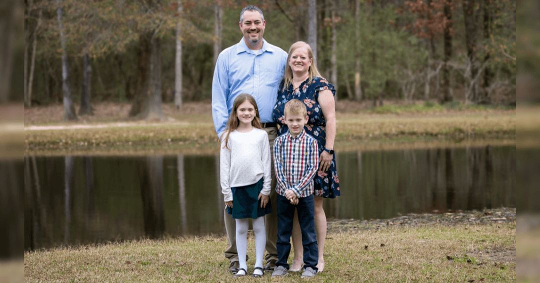 Family of four posing outdoors by a pond, with two parents standing behind a girl and boy.
