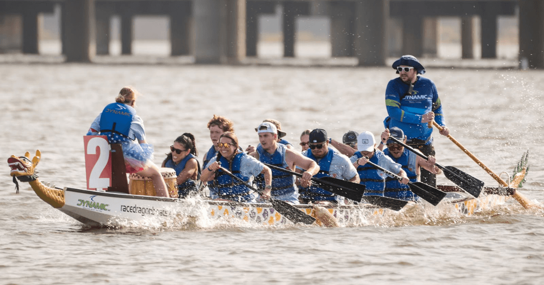 Dragon boat team paddling in unison on water, with a drummer and steersman, near a bridge.