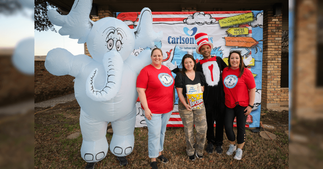 Four people in Dr. Seuss costumes pose by an inflatable elephant and Carlson Law Firm banner at an outdoor event.