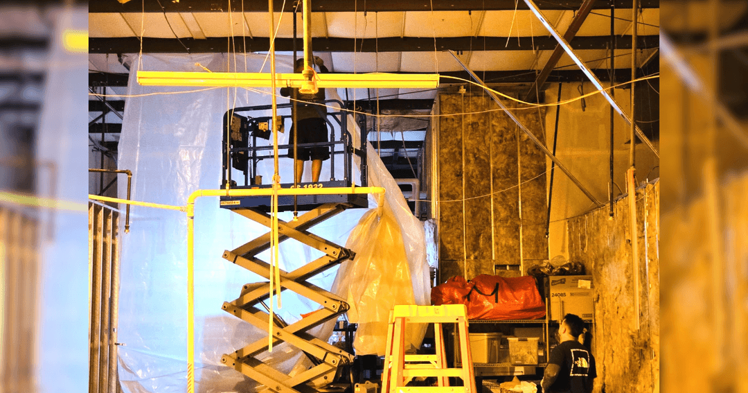 Worker on a scissor lift installs ceiling fixtures inside a building under construction with plastic sheeting.