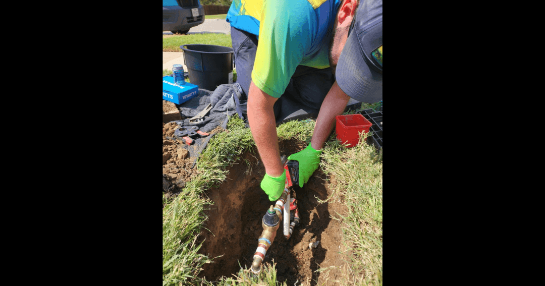 Worker in green gloves installs a water backflow valve in a dug trench in a lawn.