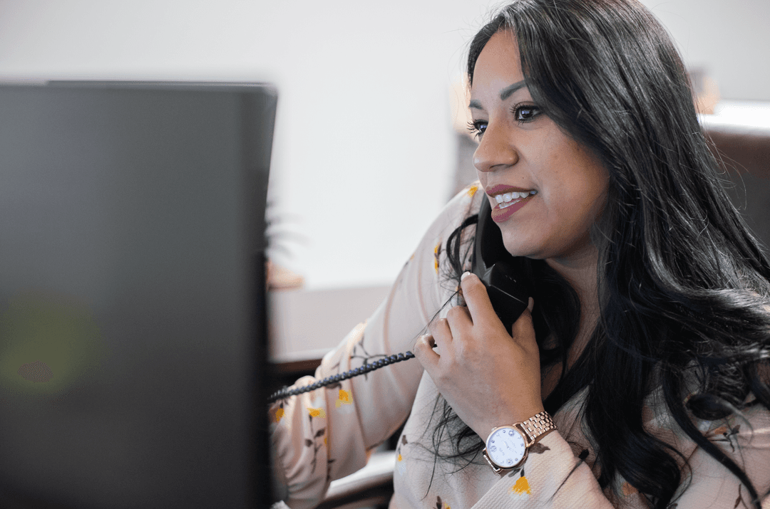 Smiling woman talking on a desk phone while looking at a computer monitor in an office.