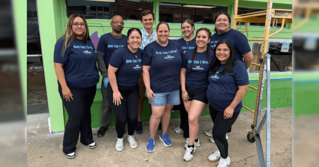 Group of nine volunteers in navy "How Can I Help?" shirts pose outside a green building near scaffolding.