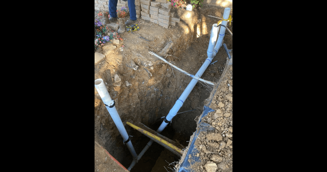 White PVC pipes descending into an excavated dirt trench during a plumbing repair, with tools and flowers nearby.