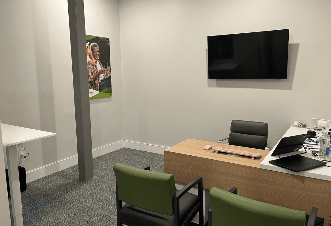 Small office with wooden desk, computer, wall-mounted TV, green guest chairs, and a framed poster.