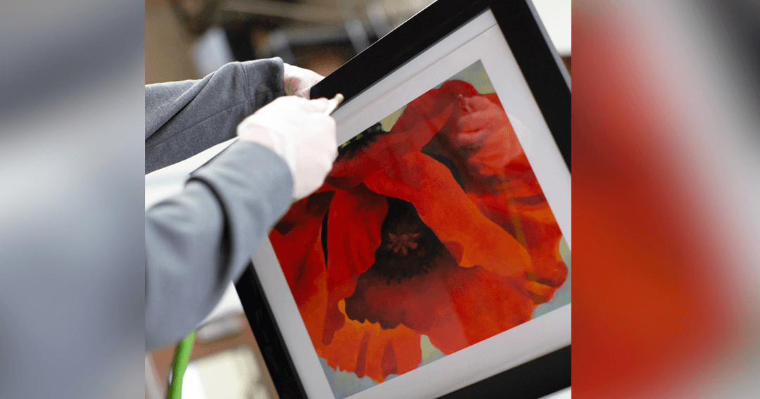 A person's hand holds a black-framed painting of a large red poppy flower.