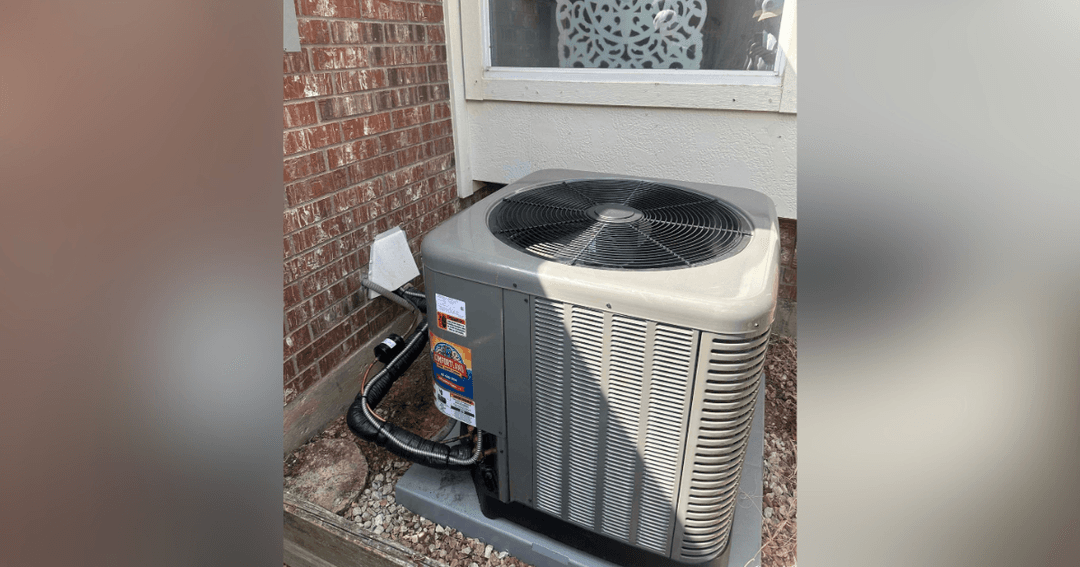 Outdoor air conditioner condenser unit installed beside a house near a brick wall and window.