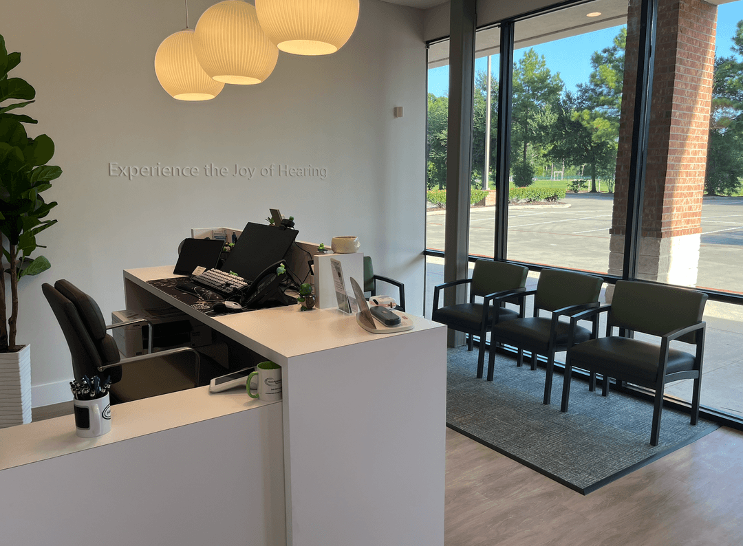 Hearing clinic reception area with desk, waiting chairs by windows, and wall text "Experience the Joy of Hearing.