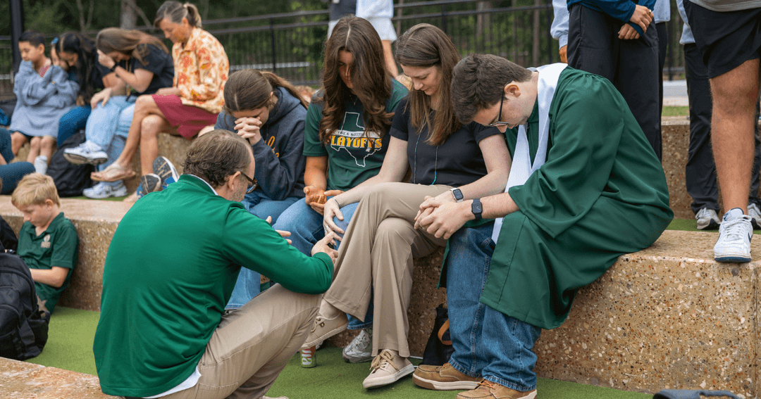 People in green attire bow their heads and hold hands in prayer while seated on outdoor stone steps.