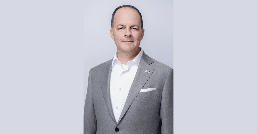 Man in a gray suit and white shirt with a lapel pin, posing against a light gray background.