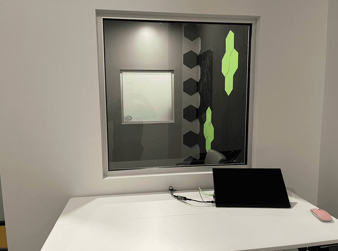 White desk with a portable monitor, cables, and pink mouse below a window into a room with green hexagonal wall panels.