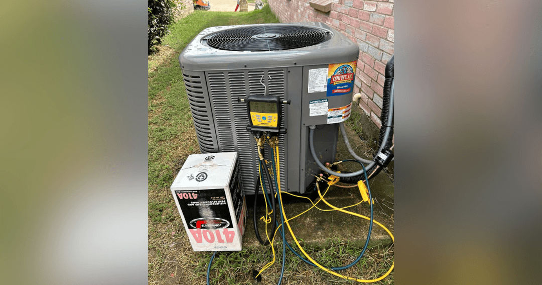 Outdoor AC condenser unit connected to digital gauges and hoses, with a box of R-410A refrigerant nearby.