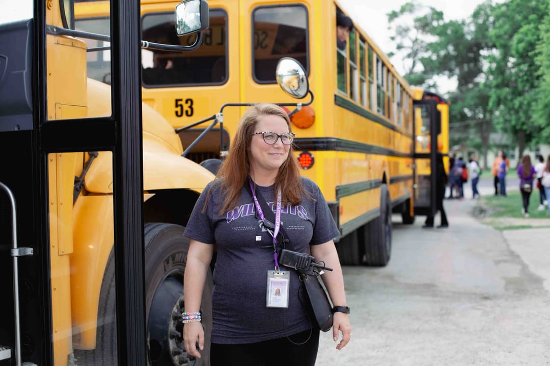 School staff member in a Wildcats shirt with ID lanyard and radio stands beside yellow school buses as students gather.