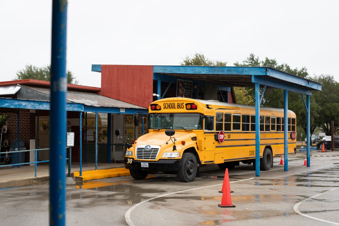 Yellow school bus parked under a covered walkway outside a school on a wet, overcast day, with orange traffic cones nearby.