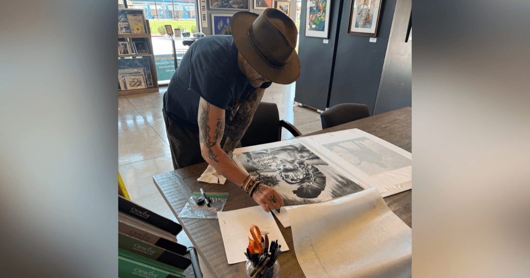 Tattooed man in a hat leans over a table examining large artwork prints in a gallery.