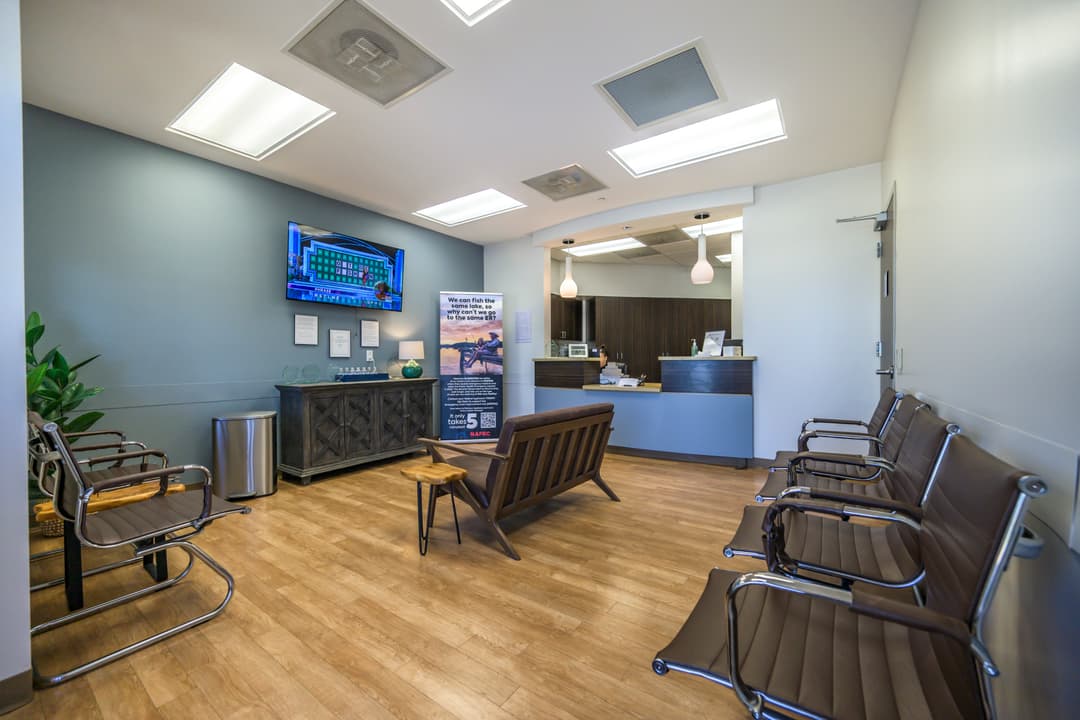 Medical office waiting room with brown chairs, wall-mounted TV, reception desk, and wood flooring.