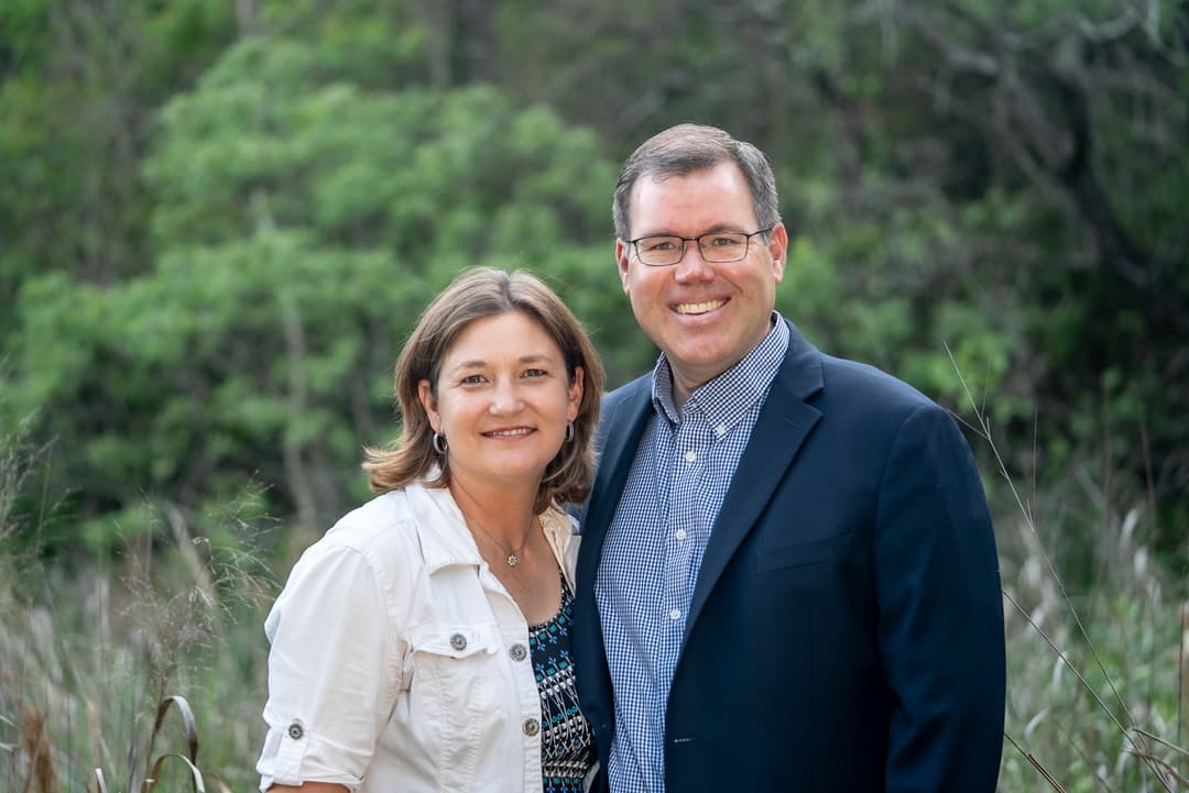 Smiling couple posing together outdoors in front of green foliage and tall grass.