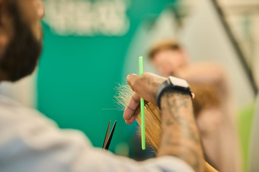 Tattooed barber cuts a client's hair with scissors and a green comb in a salon.