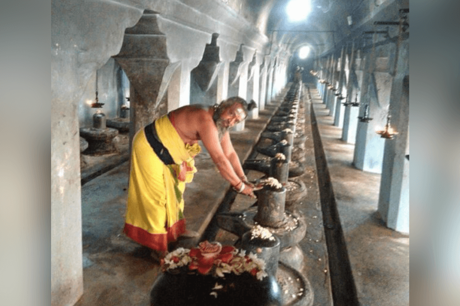 Priest in yellow robe performs rituals on a long row of stone Shiva lingams inside a temple hall.
