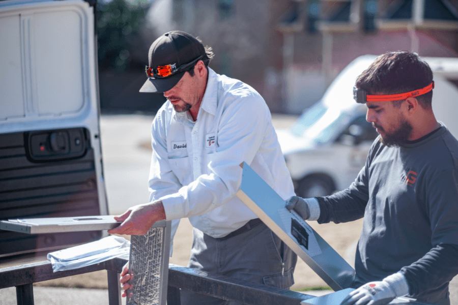 Two workers in caps handle metal ductwork materials near a work van outdoors.
