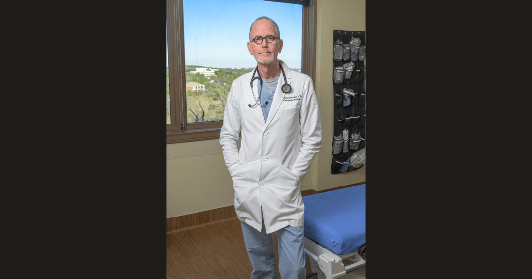 Doctor in white coat with stethoscope stands by a window in a hospital room, hands in pockets.