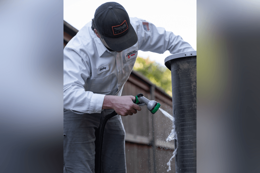 HVAC technician in uniform and cap uses a hose to spray water and clean an air conditioner condenser coil.
