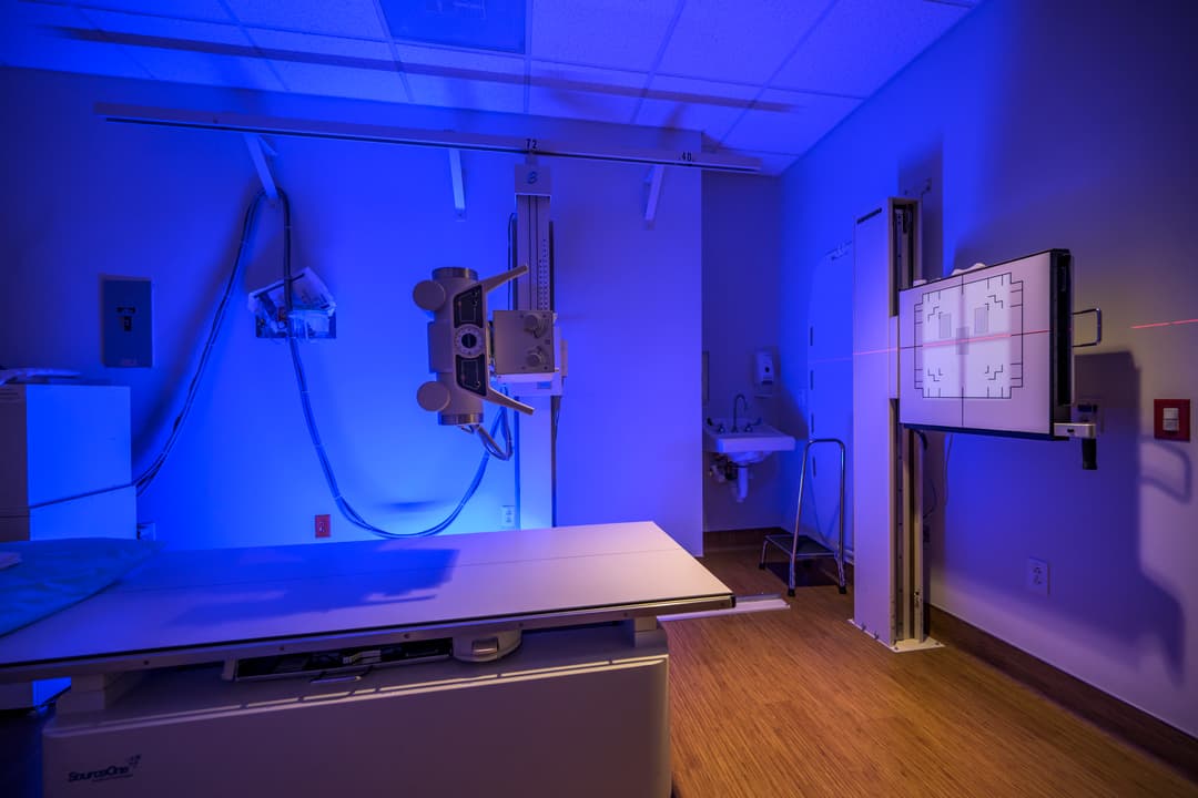 Radiology X-ray room with imaging table, ceiling-mounted tube and wall detector, lit by blue mood lighting.