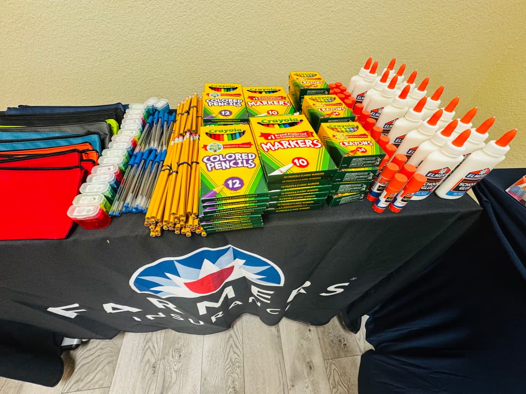 School supplies including pencils, markers, crayons and glue on a table with a Farmers Insurance cloth.