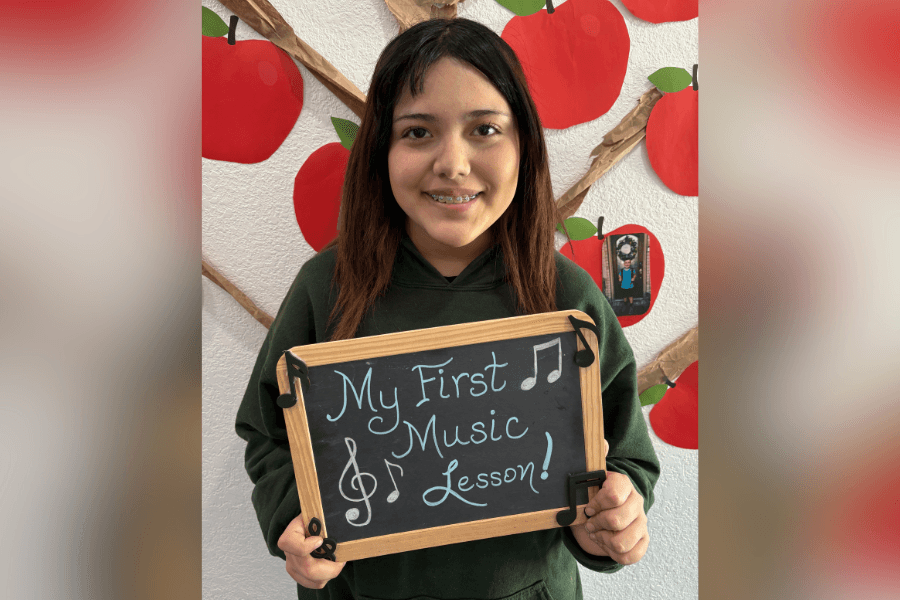 Smiling girl holds chalkboard reading "My First Music Lesson!" with music notes, in front of apple tree display.