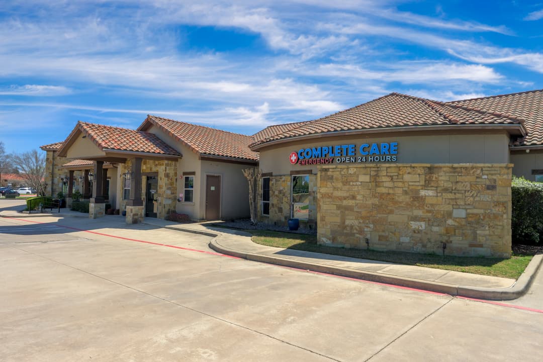 Complete Care emergency clinic, a stone building with tile roof and "Open 24 Hours" sign, under blue sky.