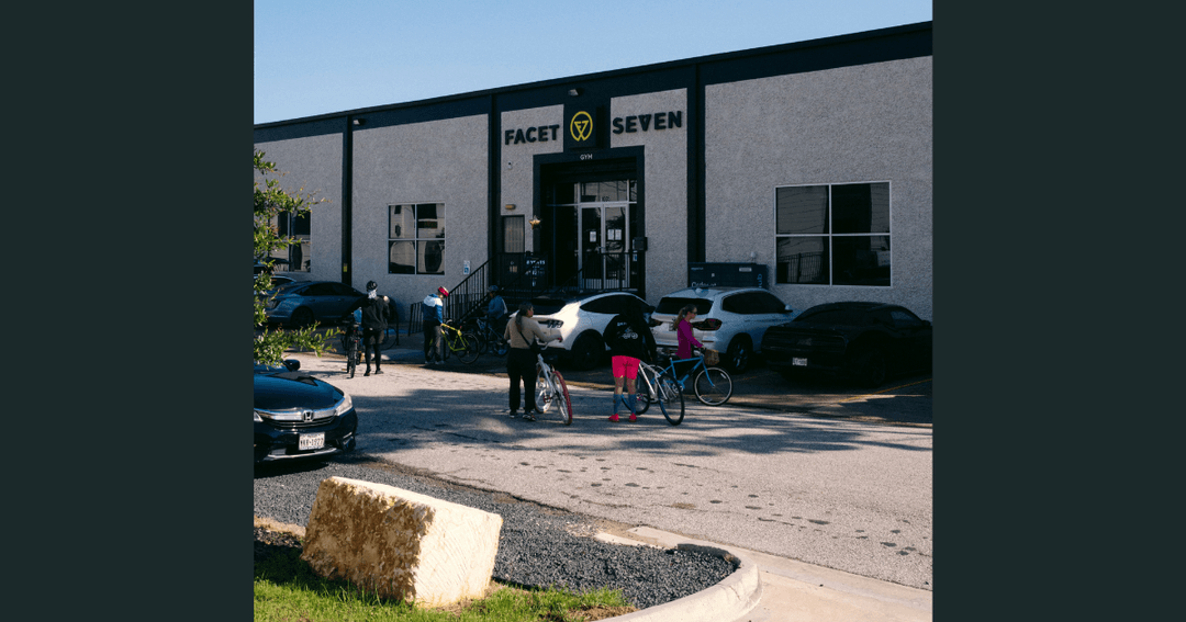 Cyclists with bikes gather outside Facet Seven gym in a parking lot with parked cars.