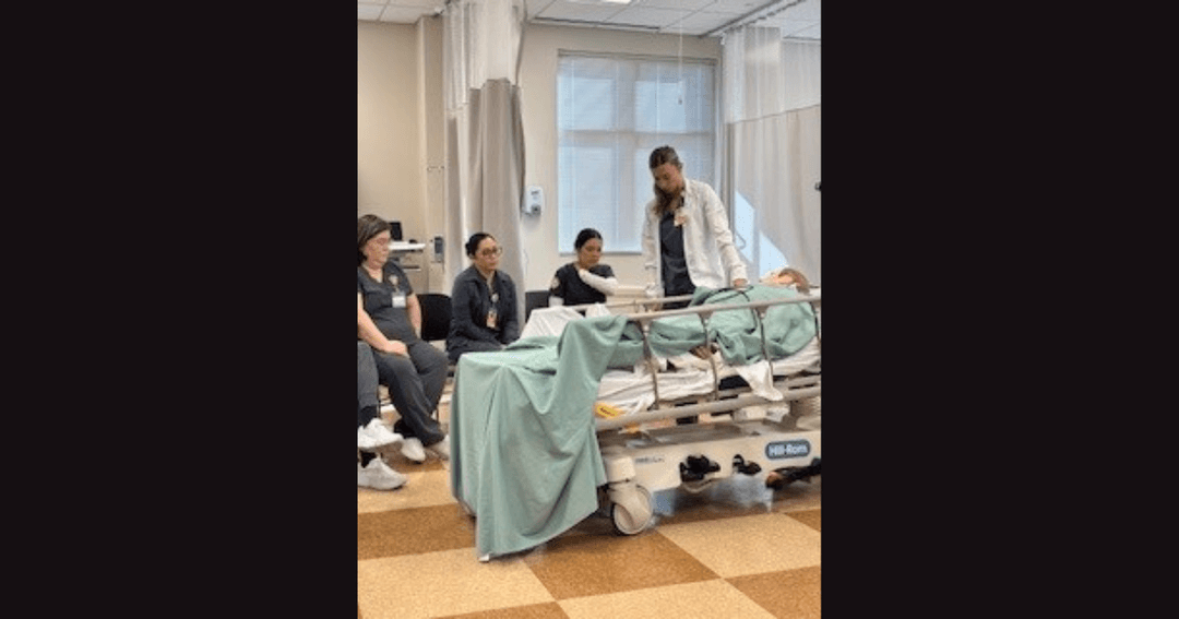 Medical instructor stands by a hospital bed with a patient as three students in scrubs observe during training.