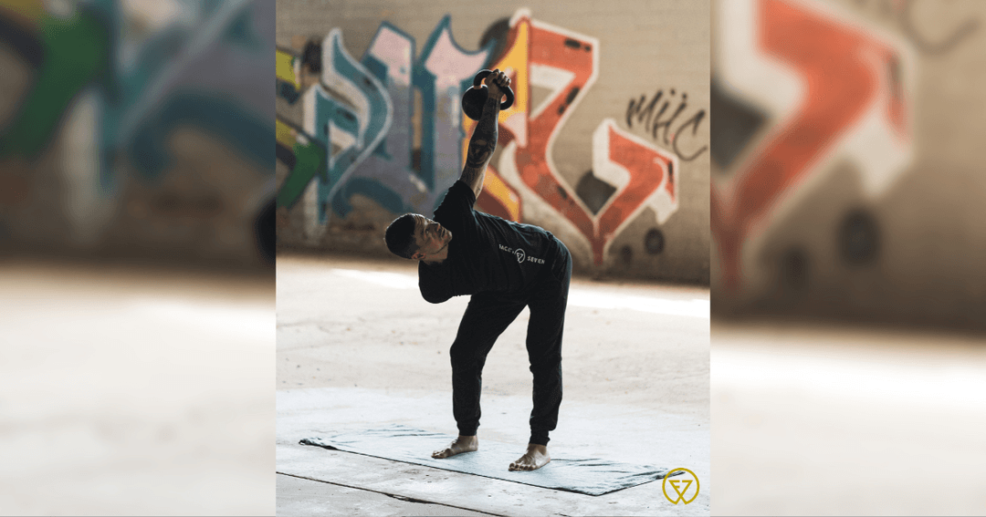 Man in black holds a kettlebell overhead doing a windmill exercise against a graffiti wall.
