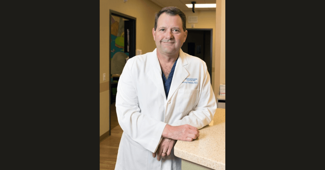 Smiling doctor in a white lab coat leaning on a counter in a medical office.