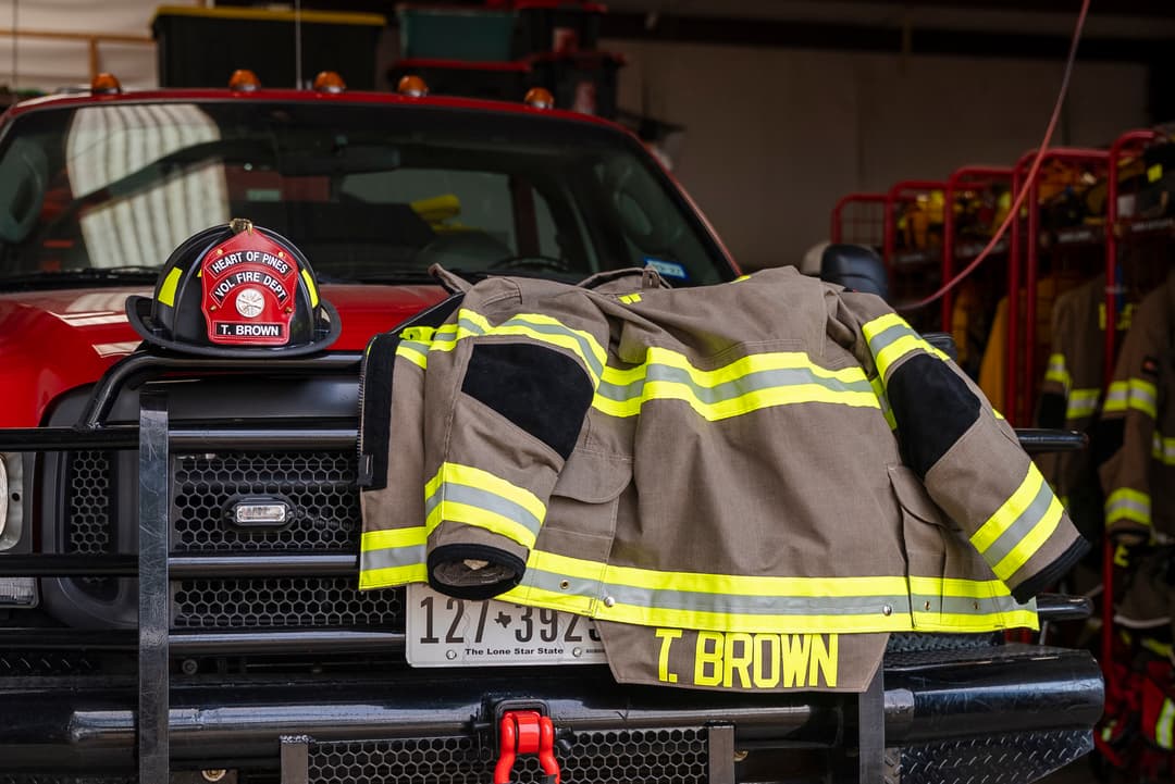 Firefighter turnout gear and helmet labeled "T. Brown, Heart of Pines Vol Fire Dept" on a red fire truck's bumper.