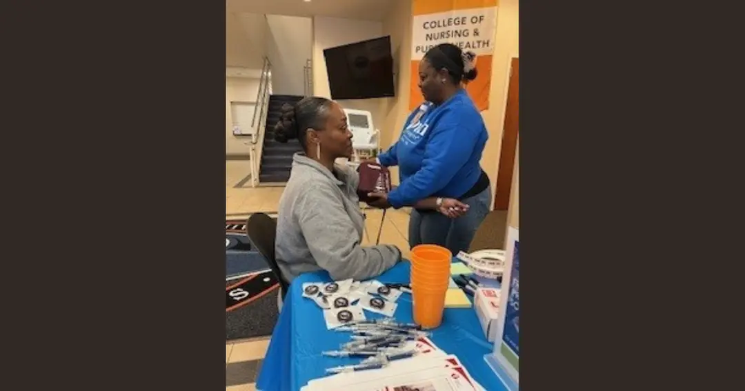 Nurse checks a seated woman's blood pressure at a College of Nursing and Public Health health fair table.
