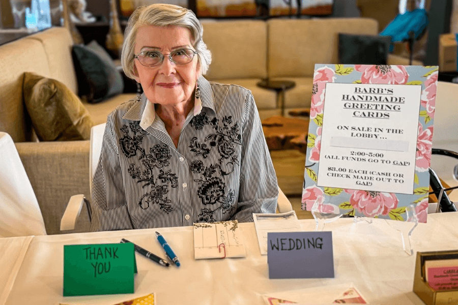 Elderly woman at table selling handmade greeting cards, with sign reading "Barb's Handmade Greeting Cards, $3 each.