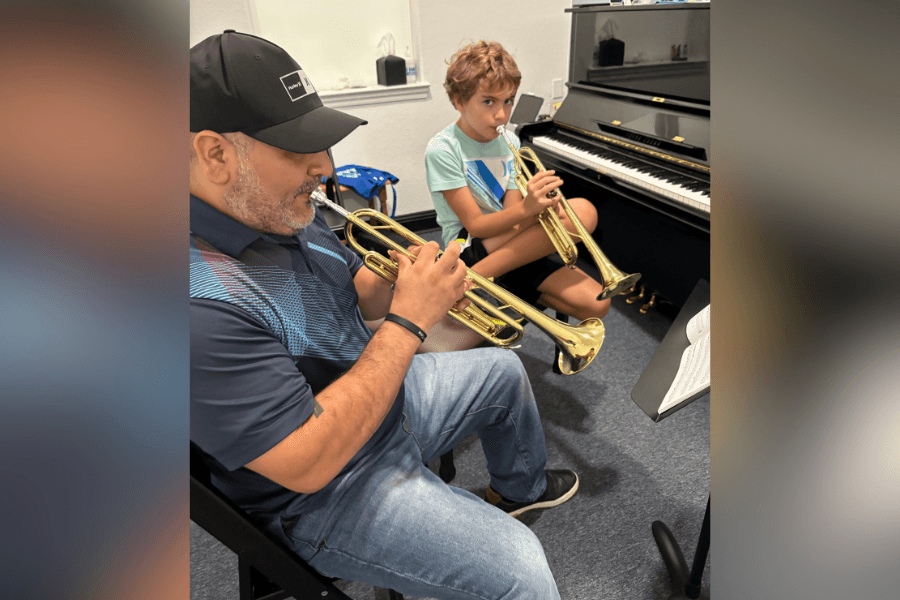 A man and a boy play trumpets together in a music room with a piano and sheet music nearby.