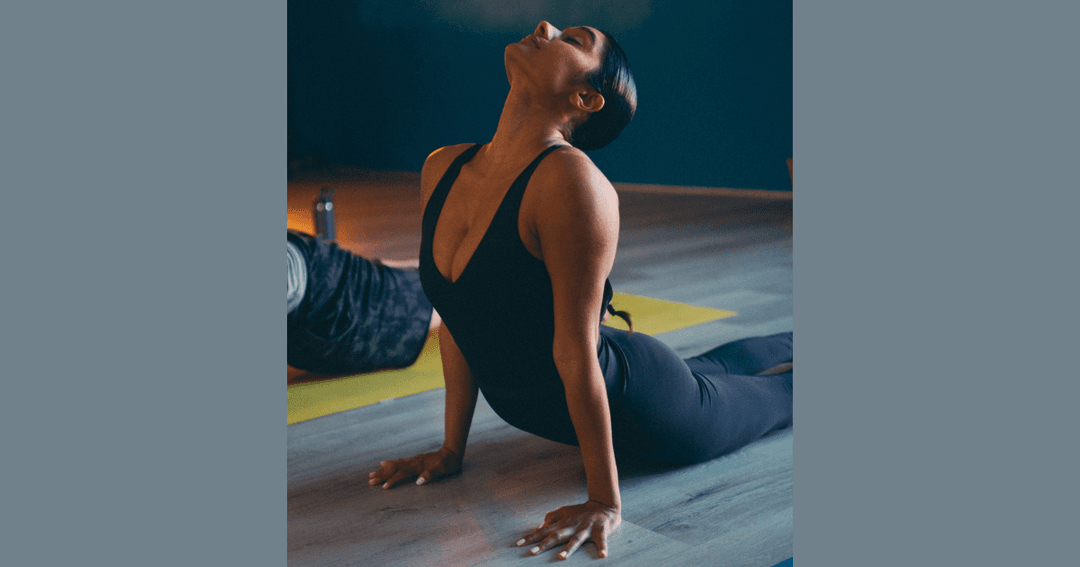 Woman in black activewear performs a yoga cobra pose on a mat in a dimly lit studio.