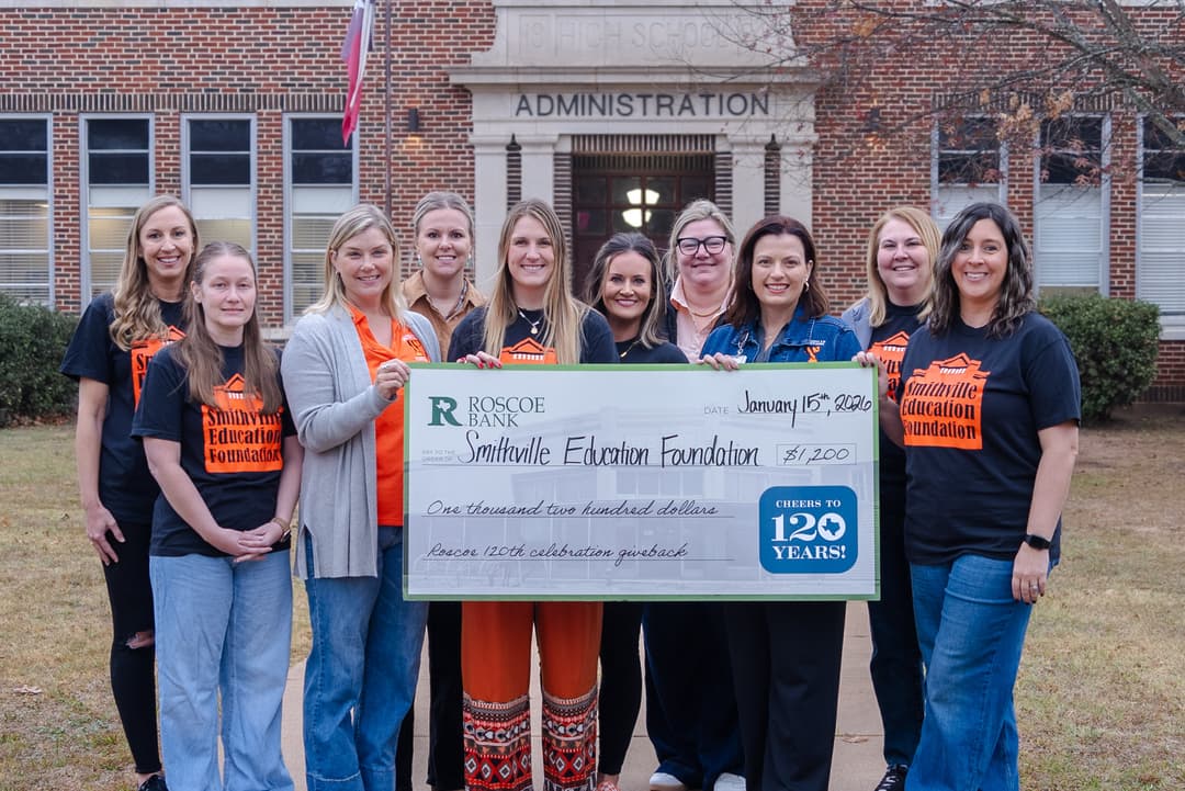 Group of women hold a large $1,200 Roscoe Bank check for Smithville Education Foundation outside a school Administration building.