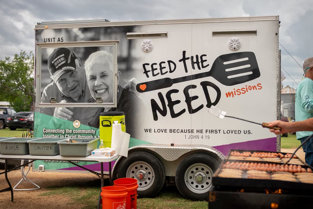 Person grills sausages beside a "Feed the Need Missions" trailer with sanitation bins at a community event.