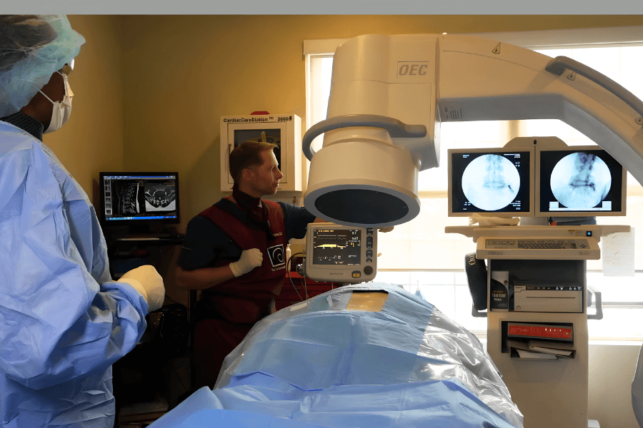 Medical staff perform a procedure using a C-arm X-ray machine, with spinal scans displayed on monitors.