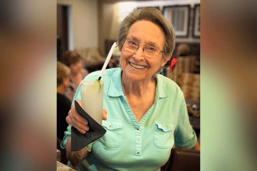 Smiling elderly woman in a teal shirt holds up a margarita with a straw at a restaurant.