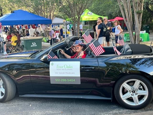 Two people in a black convertible with American flags at an outdoor event, car door reads "Four Oaks Medicare Planning.