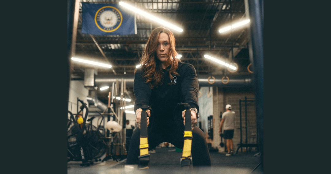 Woman in black workout gear uses suspension straps in a gym with a US Navy flag overhead.
