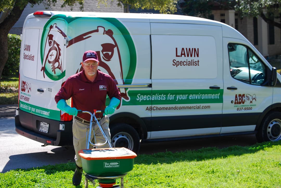 ABC lawn specialist in maroon shirt pushes a fertilizer spreader across grass in front of a company van.