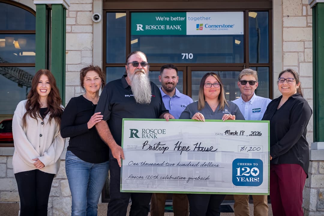 Seven people hold a giant $1,200 Roscoe Bank check for Bastrop Hope House outside the bank building.