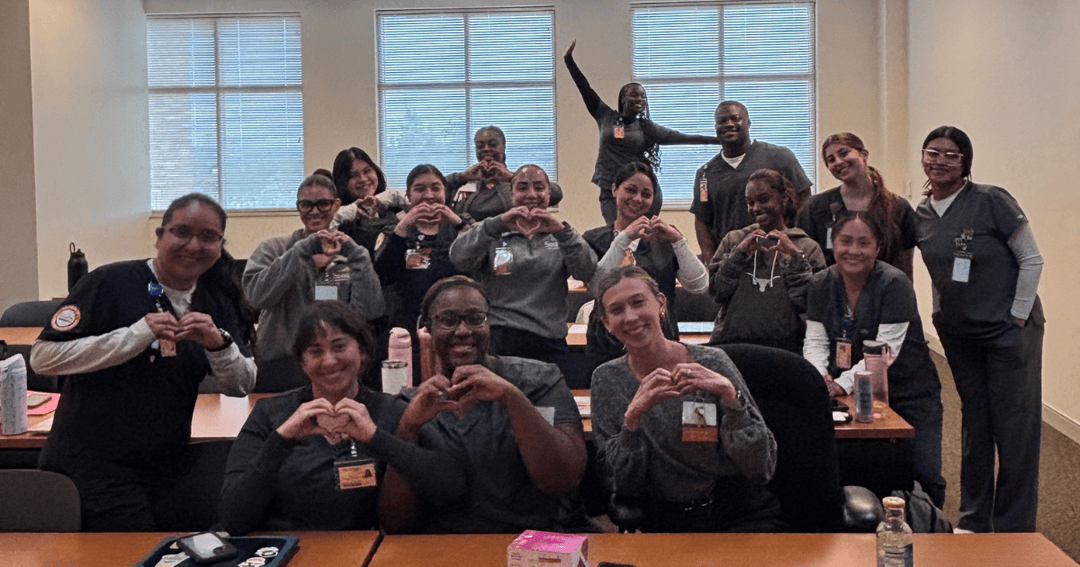 Group of students in scrubs making heart shapes with their hands in a classroom.