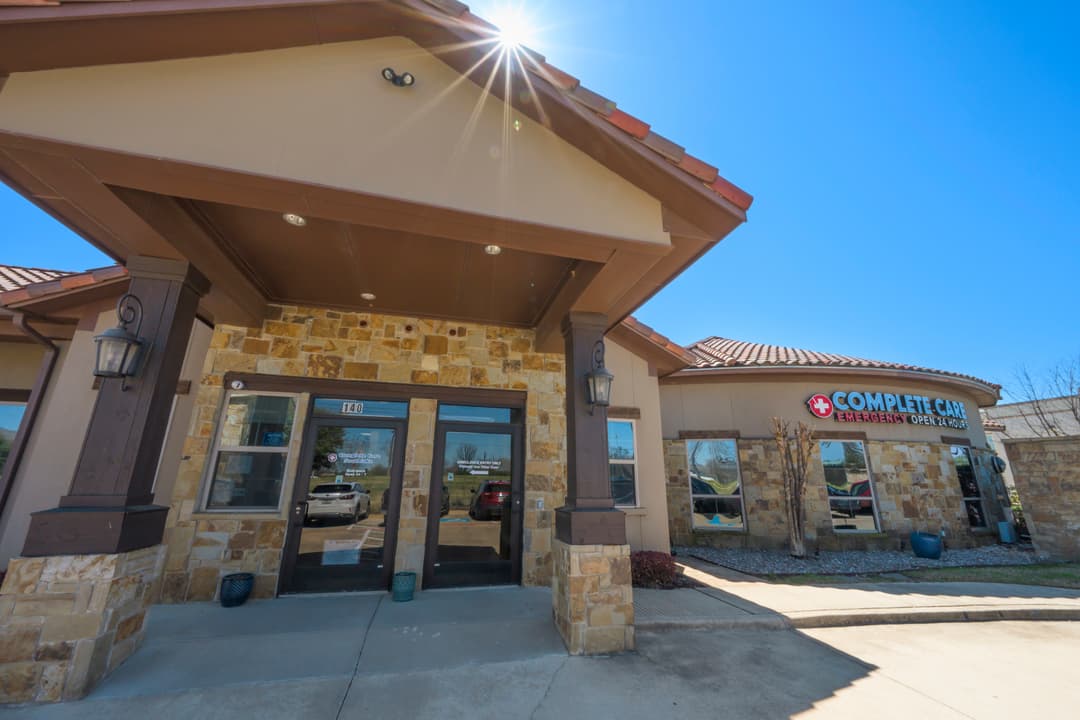 Exterior entrance of Complete Care Emergency, a 24-hour facility with stone walls and covered doorway under blue sky.