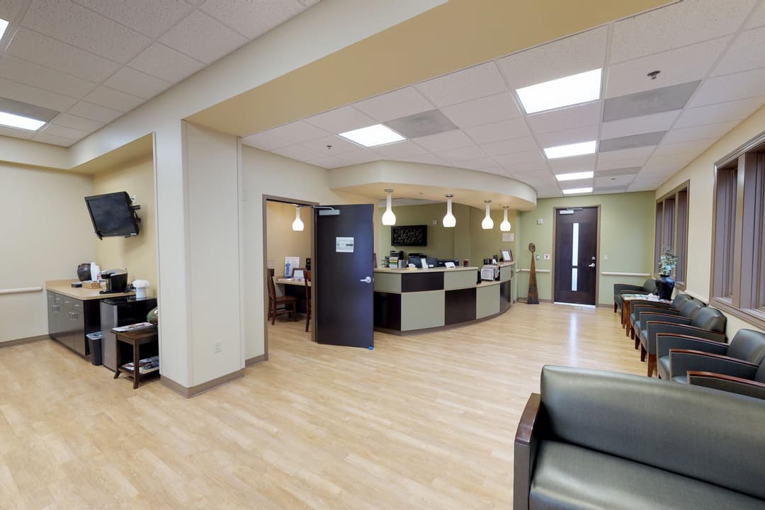 Medical office waiting room with reception desk, green leather chairs, wall-mounted TV, coffee station and wood floors.